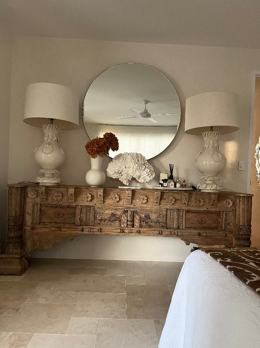 Decorative wooden console table with round mirror, lamps, and decorative items in a room.