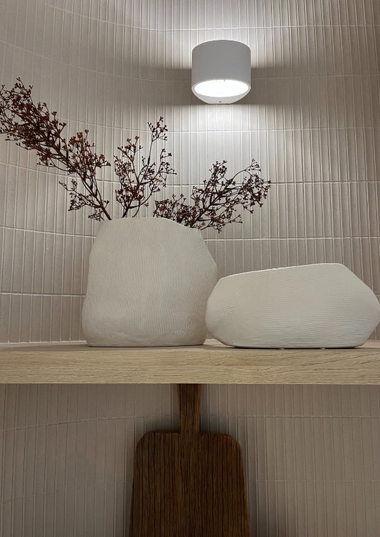 Two white vases on a wooden shelf with a light fixture above against a tiled wall.