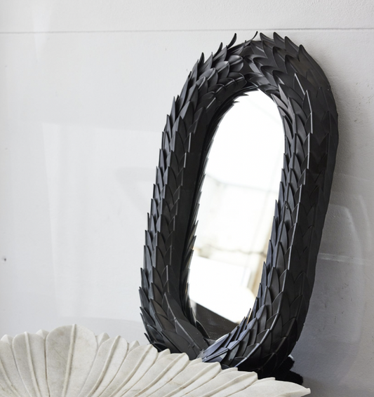 A handcrafted wreath mirror with a black frame made of leaf-like elements, hanging on a white wall.