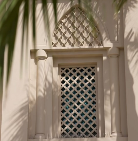Decorative window with lattice design on a building facade, surrounded by palm leaves.