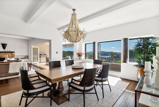 Dining room with wooden table and chairs, chandelier, and large windows overlooking a scenic view. Home styling Bungalow Interiors Gold Coast