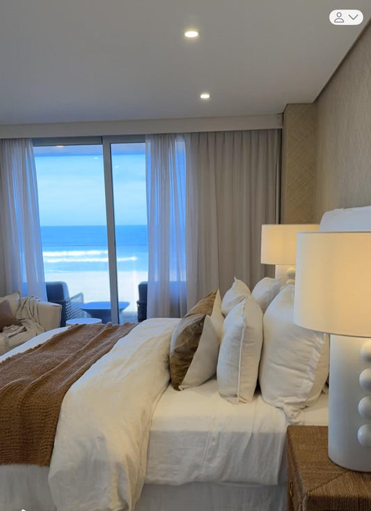 Bedroom with a large window view of the ocean, featuring a bed with white and brown bedding and two lamps. Bungalow Interiors