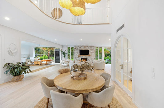 Modern living room with a round wooden table and chairs, featuring a fireplace and large windows.  Bungalow Interiors Gold Coast, coastal interior design styling
